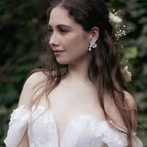 A bride in an off-the-shoulder lace dress wearing the Titania sculpted metal floral petal and pearl drop earrings in a forest setting.